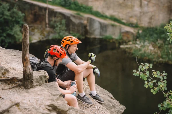 Два самца туристов в защитных шлемах с рюкзаками отдыхают с водой и яблоком на скалистом утесе — стоковое фото
