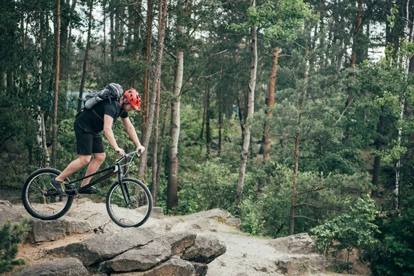Вид сбоку на экстремального велосипедиста в защитном шлеме, делающего трюк на горном велосипеде в лесу — стоковое фото