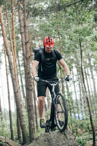 Vue de face du cycliste extrême masculin en casque de protection faisant cascade sur le vélo de montagne en forêt — Photo de stock