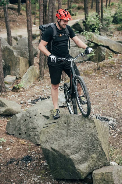 Souriant mâle extrême cycliste en casque de protection debout avec vélo de montagne sur pierre dans la forêt — Photo de stock