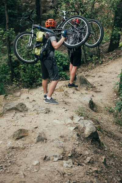 Rear view of two male extreme cyclists in helmets carrying mountain bikes in forest — Stock Photo