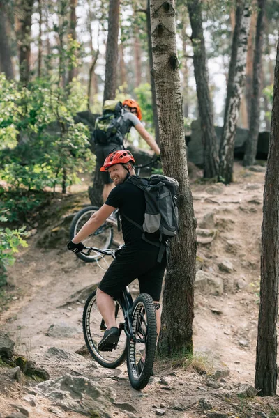 Sonriente ciclista extremo macho montando en bicicleta de montaña con amigo en el bosque - foto de stock
