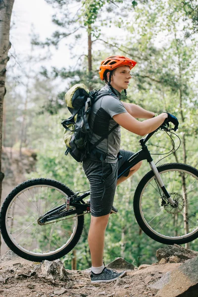 Vista lateral do ciclista masculino no capacete com mochila de pé com mountain bike na floresta — Fotografia de Stock