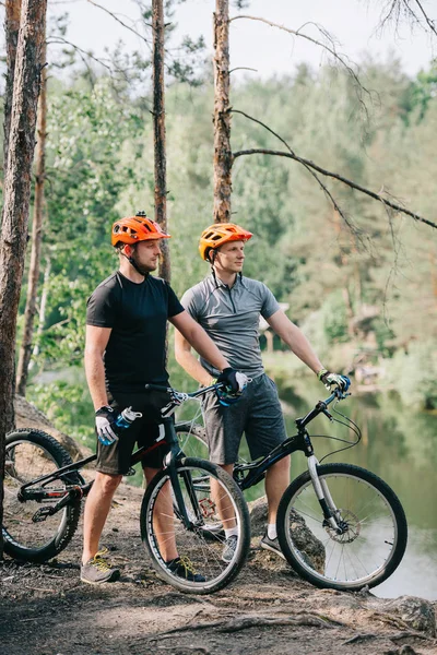 Male trial bikers in helmets with mountain cycles resting with sport bottles of water near river in forest — Stock Photo