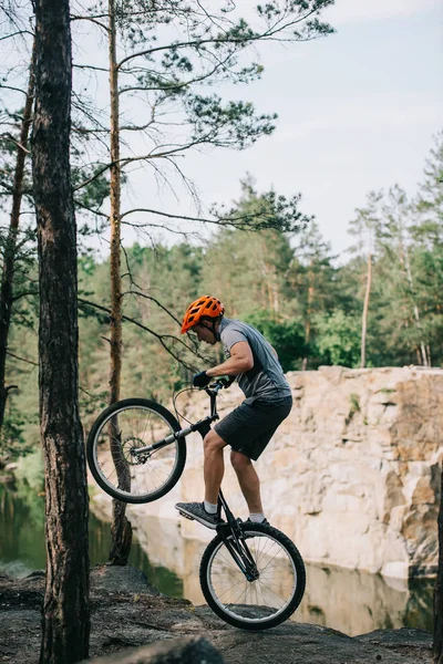 Visão lateral do ciclista extremo no capacete de proteção balanceamento na roda traseira da bicicleta de montanha na floresta — Fotografia de Stock