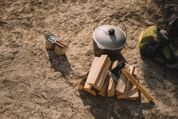 Close up view of canned food, forks, cauldron, axe, logs and backpacks on ground — Stock Photo