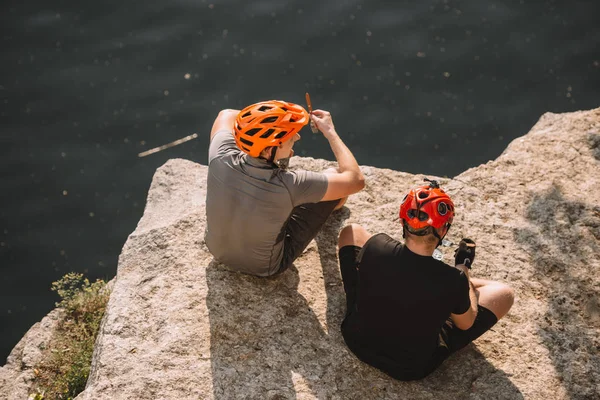 Male hikers in protective helmets resting and eating canned food on rocky cliff over river — Stock Photo