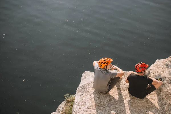 Rückansicht männlicher Reisender in Schutzhelmen, die Konserven auf felsiger Klippe über dem Fluss essen — Stockfoto