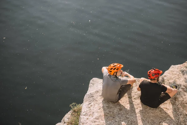 Rear view of male travelers in protective helmets eating canned food on rocky cliff over river — Stock Photo