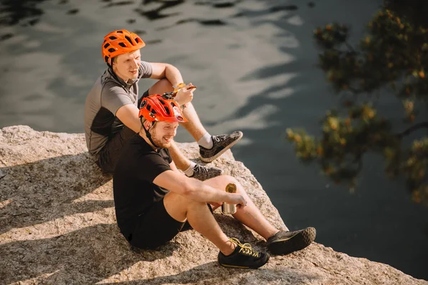 Lächelnde männliche Reisende in Schutzhelmen essen Konserven auf felsiger Klippe über dem Fluss — Stockfoto