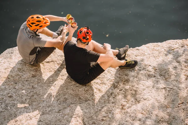 High angle view of male travelers in protective helmets clinking by cans on rocky cliff over river — Stock Photo