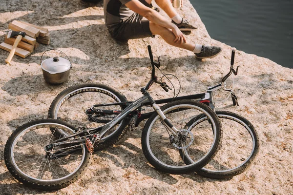 Immagine ritagliata del viaggiatore maschio seduto a terra vicino a biciclette, calderoni, asce e tronchi vicino al fiume — Foto stock