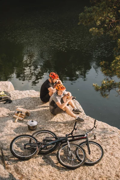 Two trial bikers resting near logs, cauldron and mountain cycles on rocky cliff over river — Stock Photo