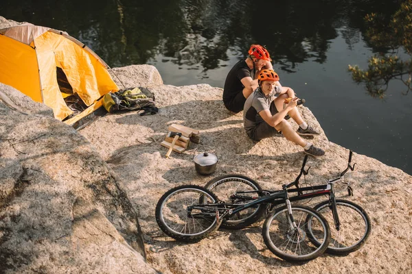 Trial bikers resting near tent and cycles on rocky cliff over river — Stock Photo