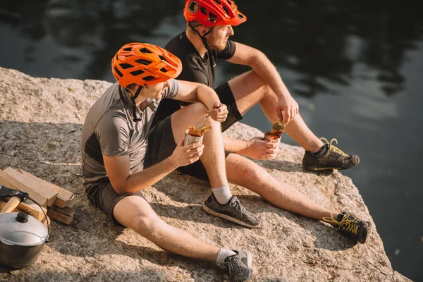 Zwei männliche Reisende essen Konserven in der Nähe von Baumstämmen, Axt und Kessel auf felsigen Klippen über dem Fluss — Stockfoto