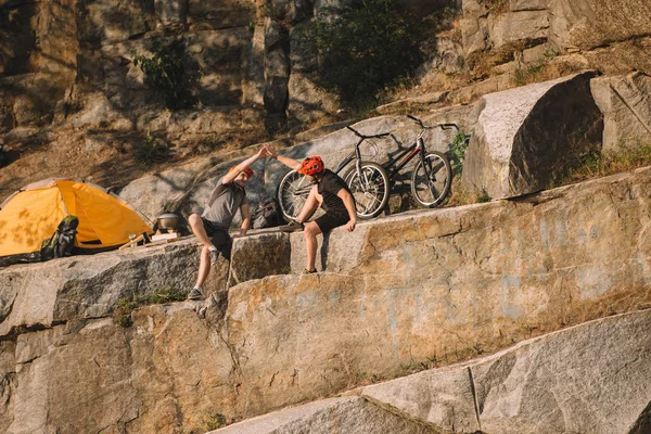 Trial bikers resting and giving high five to each other near tent and mountain cycles on rocky cliff — Stock Photo