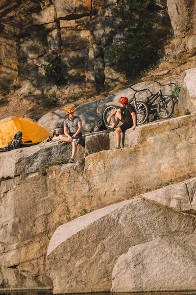 Dos viajeros masculinos en cascos de protección que descansan cerca de la tienda de campaña y bicicletas en un acantilado rocoso - foto de stock