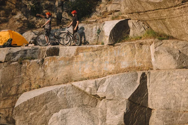 Male traveler in protective helmet standing with mountain bike and pointing by finger to friend near tent on rocky cliff — Stock Photo