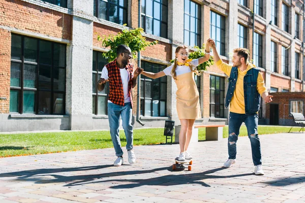 Hombres multiétnicos ayudando a las jóvenes a patinar en longboard en la calle - foto de stock