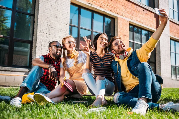 Sonrientes amigos multiétnicos tomando selfie en el teléfono inteligente mientras descansan en el césped verde juntos - foto de stock