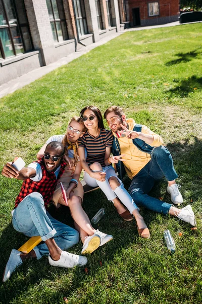 Multiethnic friends taking selfie on smartphone while resting on green lawn together — Stock Photo