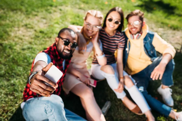 Foco seletivo de amigos multiétnicos tomando selfie no smartphone enquanto descansa no gramado verde no parque — Fotografia de Stock