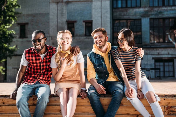 Parejas alegres multiculturales sentados en parapeto de madera juntos - foto de stock