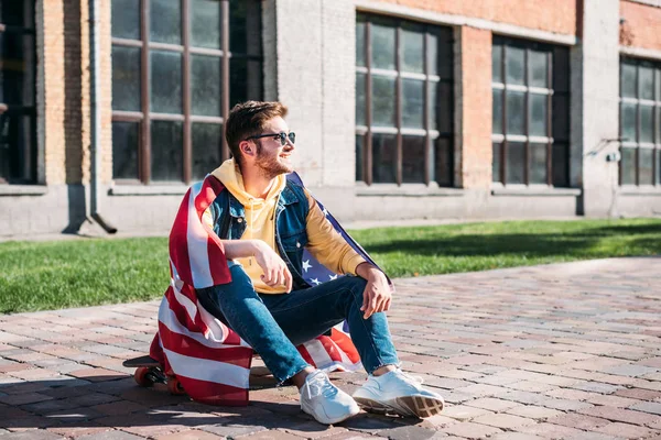 Jeune homme souriant avec drapeau américain assis sur longboard dans la rue — Photo de stock