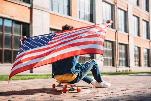 Back view of man with american flag sitting on longboard on street — Stock Photo