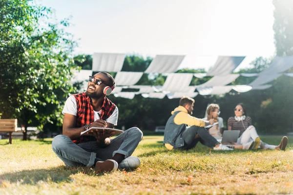Foco seletivo do homem americano africano em fones de ouvido com notebook descansando na grama verde com amigos multirraciais com laptop atrás — Fotografia de Stock