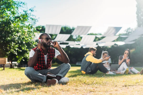 Enfoque selectivo del hombre afroamericano en auriculares con portátil descansando sobre hierba verde con amigos multirraciales con ordenador portátil detrás - foto de stock