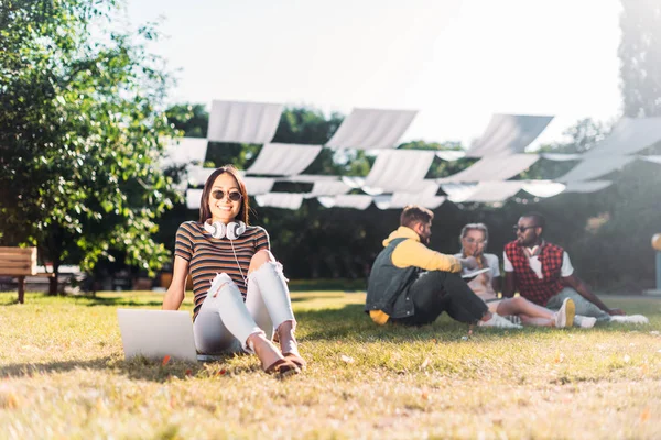 Selective focus of young asian woman with laptop and multiracial friends behind resting on green grass in park — Stock Photo