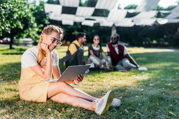 Selective focus of young smiling woman with laptop and multiracial friends behind resting on green grass in park — Stock Photo