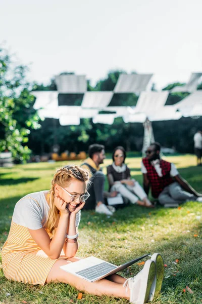 Enfoque selectivo de la joven mujer sonriente con el ordenador portátil y amigos multirraciales detrás de descansar en la hierba verde en el parque - foto de stock
