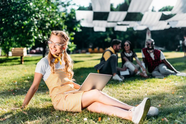 Foco seletivo de jovem mulher sorridente com laptop e amigos multirraciais por trás descansando na grama verde no parque — Fotografia de Stock