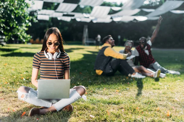 Selective focus of smiling asian woman using laptop and multiracial friends behind resting on green grass in park — Stock Photo