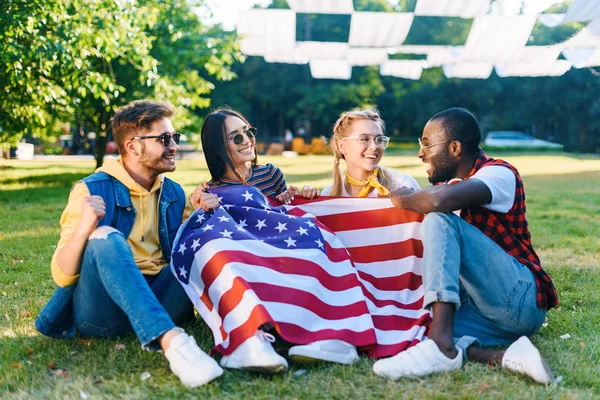 Amigos sonrientes multirraciales con bandera americana sentados sobre hierba verde en el parque - foto de stock