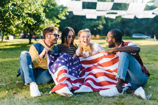 Многорасовые счастливые друзья с американским флагом сидят на зеленой траве в парке — стоковое фото