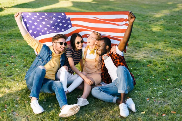 Multirracial amigos alegres com bandeira americana sentado na grama verde no parque — Fotografia de Stock