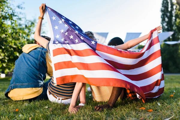 Vista trasera de amigos multirraciales con bandera americana sentados sobre hierba verde en el parque - foto de stock