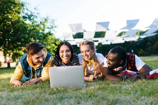 Retrato de jóvenes amigos interracial con ordenador portátil descansando en el césped verde en el parque - foto de stock