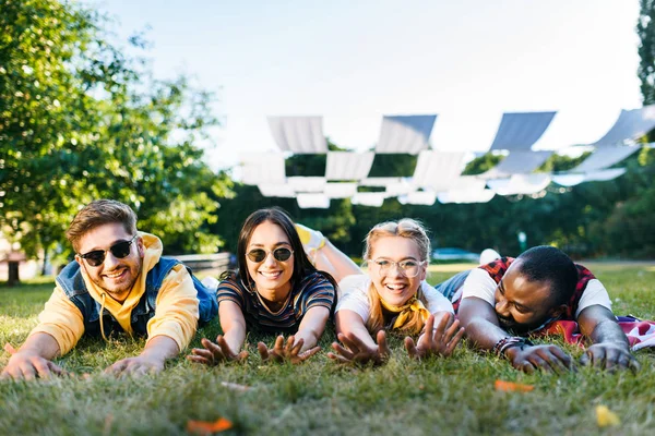 Retrato de alegres amigos jóvenes interracial tumbados sobre hierba verde en el parque en el día de verano - foto de stock