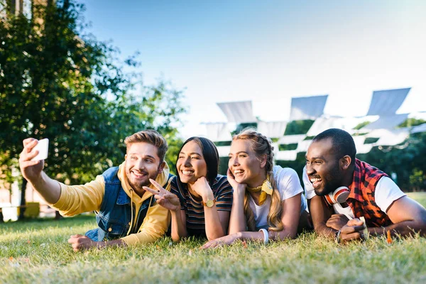 Portrait d'amis gais multiethniques prenant selfie sur smartphone tout en se reposant sur l'herbe verte dans le parc — Photo de stock