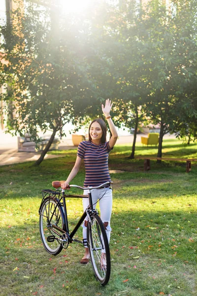Молодая азиатка с ретро-велосипедом приветствует кого-то в парке — стоковое фото