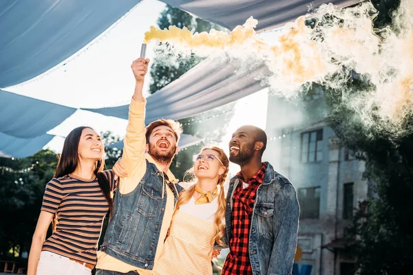 Interracial cheerful young friends with colorful smoke bomb in park — Stock Photo