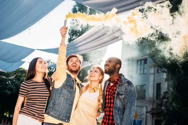 Interracial jovens amigos alegres com bomba de fumaça colorida no parque — Fotografia de Stock