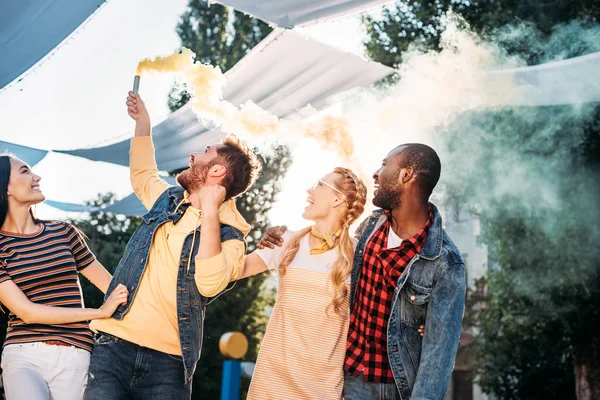Interracial jovens amigos alegres com bomba de fumaça colorida no parque — Fotografia de Stock