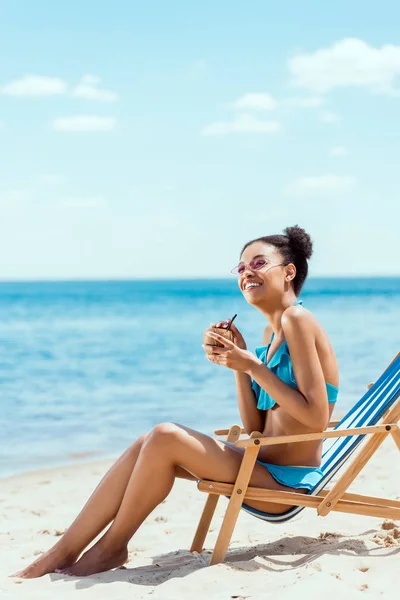 Smiling african american woman in bikini and sunglasses drinking cocktail in coconut shell while sitting on deck chair in front of sea — Stock Photo