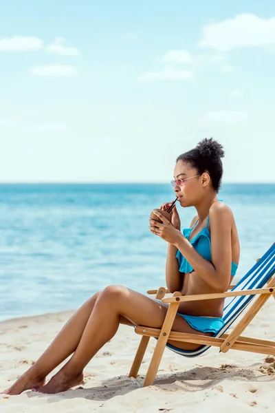 Молода жінка в бікіні і сонцезахисні окуляри, пити коктейль в шкаралупи кокосового горіха, сидячи на шезлонгу березі моря — стокове фото