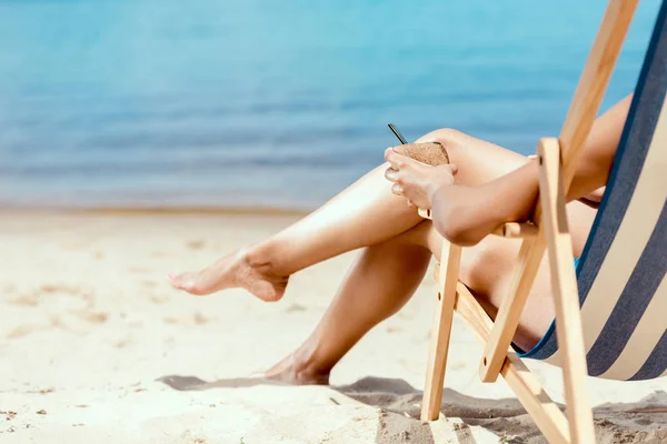 Imagen recortada de la mujer sosteniendo cóctel en cáscara de coco y la colocación en la silla de cubierta en la playa de arena - foto de stock
